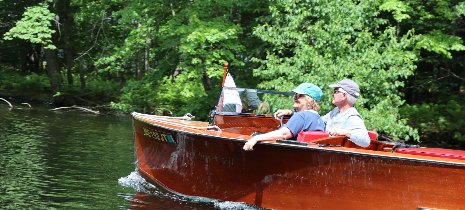 Two club members smiling as they drive a mahogany runabout up the Songo River