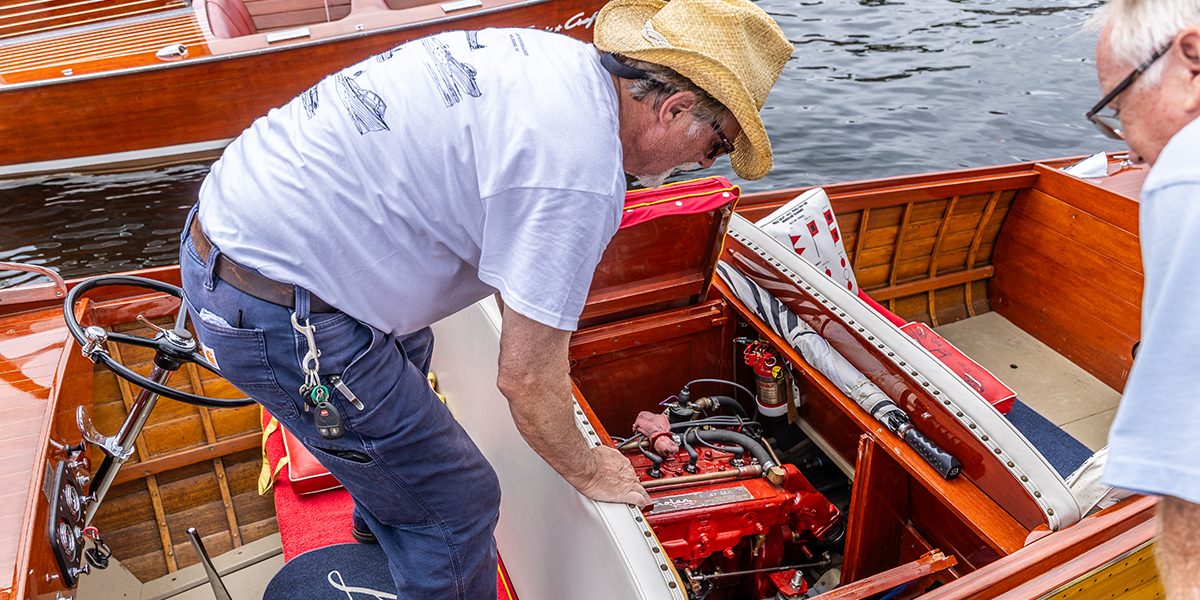A club member leans into a classic runabout to work on its engine while a friend looks on